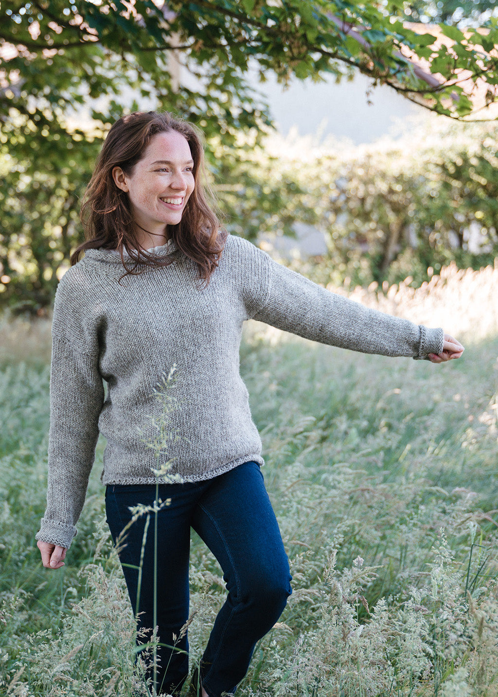 A woman smiling outdoors in a grassy field, wearing a light grey-brown knit jumper and dark blue jeans.