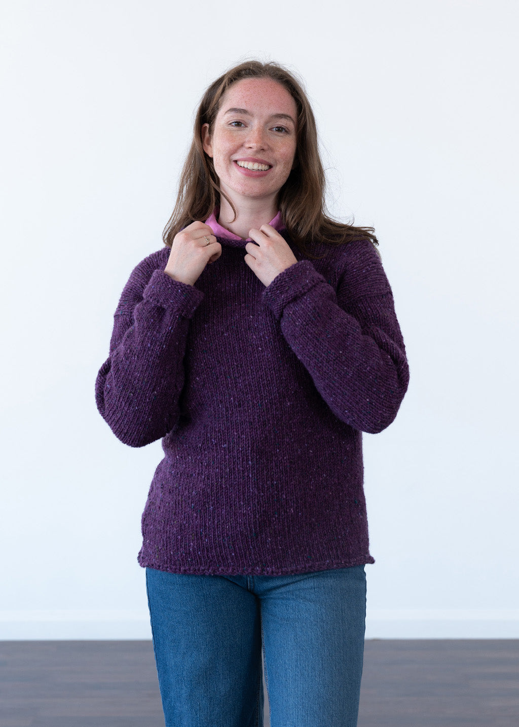 A smiling woman wearing a chunky knit purple jumper and blue jeans, holding the collar of the sweater. The background is a clean white studio wall with a dark floor visible at the bottom.