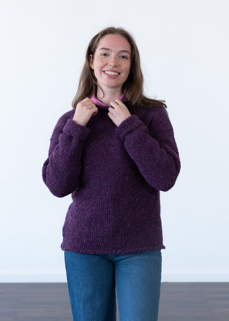 A smiling woman wearing a chunky knit purple jumper and blue jeans, holding the collar of the sweater. The background is a clean white studio wall with a dark floor visible at the bottom.