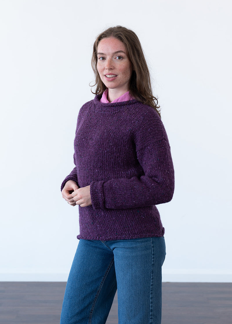 A woman wearing a chunky knit purple sweater over a pink collared shirt and blue jeans, standing against a plain white wall.