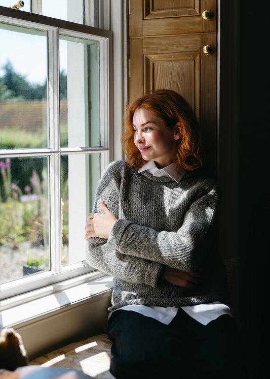 A woman with red hair sits by a window wearing a grey knit jumper over a white collared shirt and dark trousers, with warm sunlight and wooden shutters in the background.
