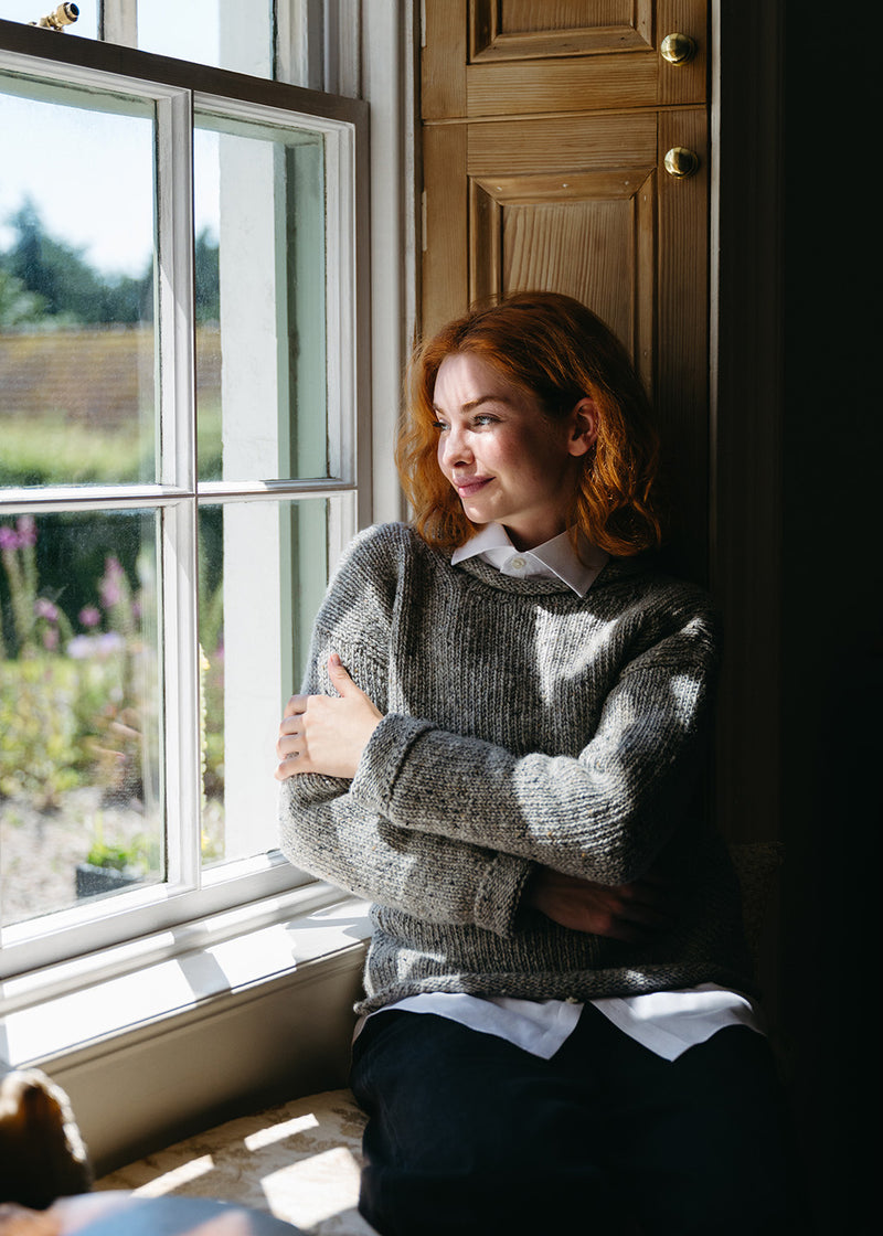 A woman with red hair sits by a window wearing a grey knit jumper over a white collared shirt and dark trousers, with warm sunlight and wooden shutters in the background.