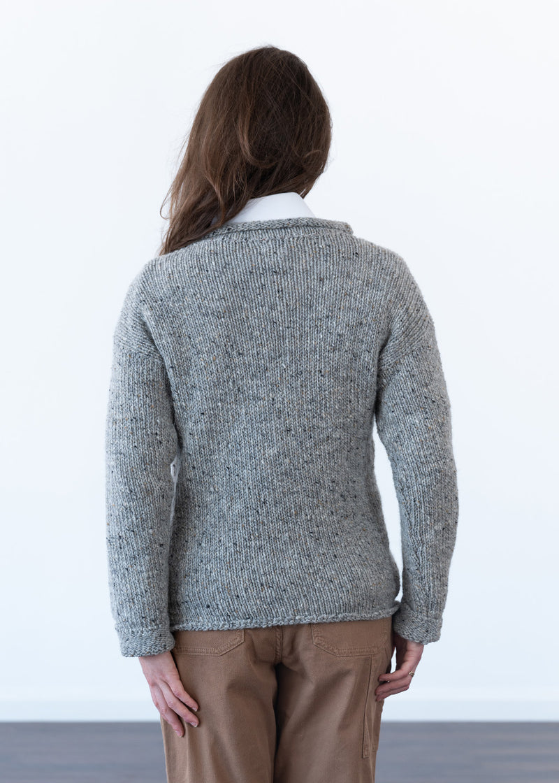 A woman viewed from behind wearing a textured grey knit jumper over a white collared shirt and brown trousers, standing against a plain white wall.