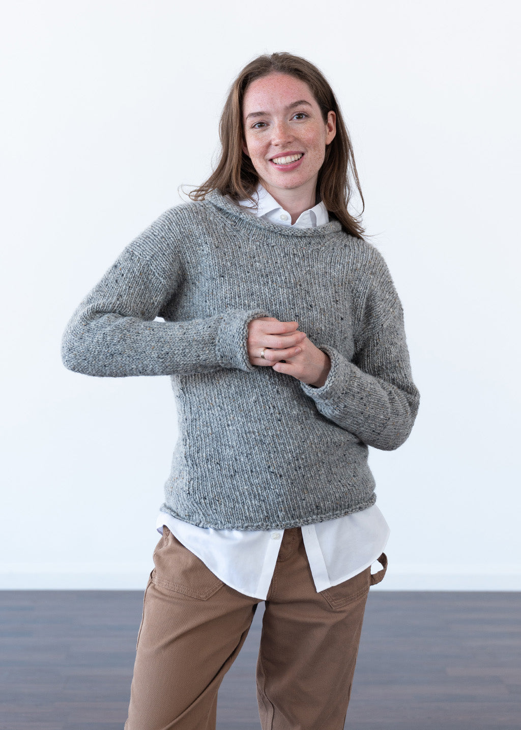 A woman modeling a chunky knit grey flecked wool jumper over a white collared shirt and brown trousers, standing against a clean white studio background.