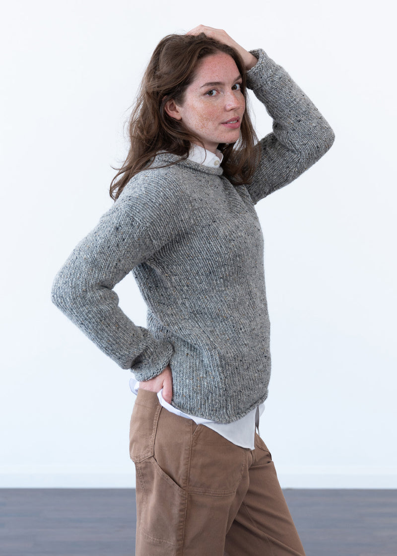 A woman modeling a chunky gray knit sweater with a rolled crew neck, paired with brown pants and a white shirt, set against a minimalist white studio background.