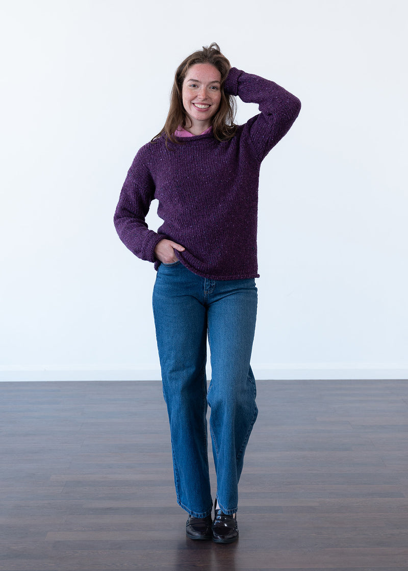 A woman modeling a chunky purple knit jumper and blue jeans against a clean white studio background.