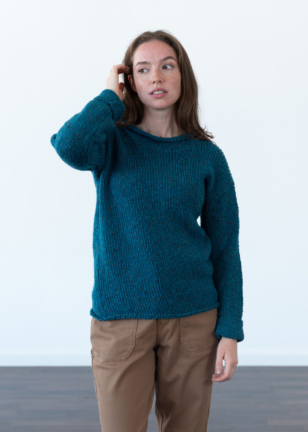 A woman wearing a chunky knit teal jumper and brown cargo trousers, standing against a plain white wall.