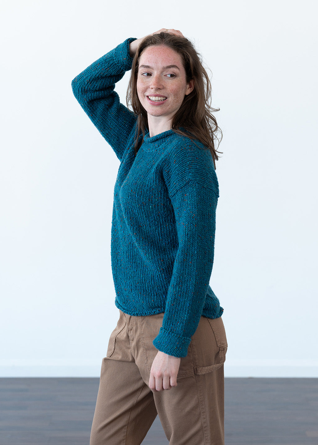 A woman wearing a teal blue chunky knit sweater and brown pants, posing in a minimalist studio setting with a white wall and dark floor.