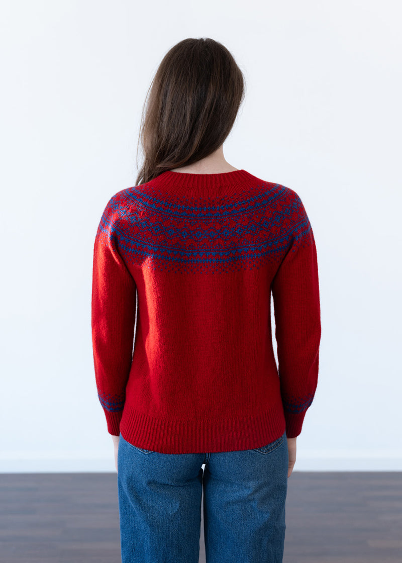 A woman viewed from behind wearing a red knit sweater with a blue Fair Isle yoke pattern, paired with blue jeans, against a plain white wall.