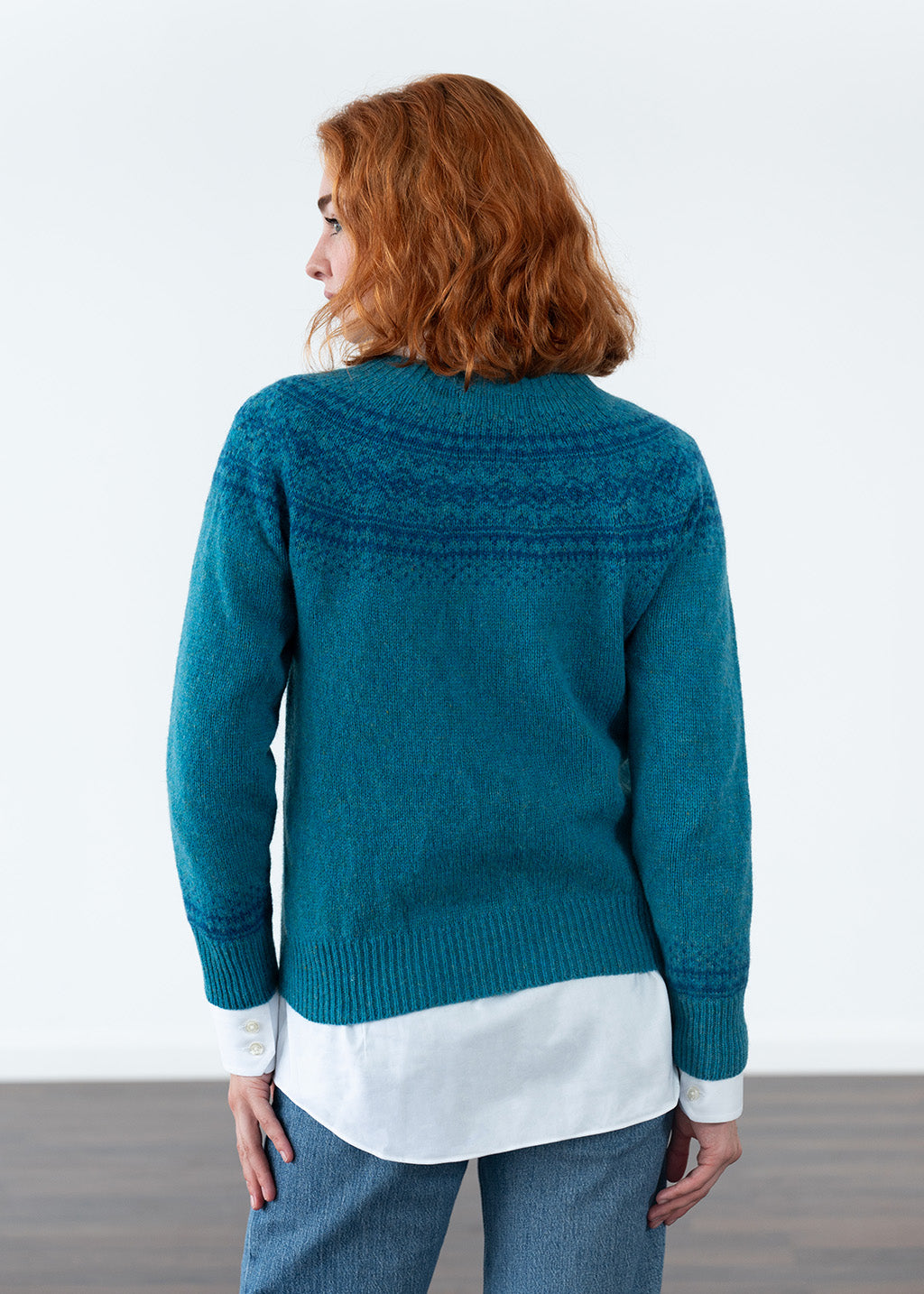 A woman with red hair is shown from the back wearing a teal blue fair isle knit sweater over a white shirt and blue jeans, set against a plain white studio background.