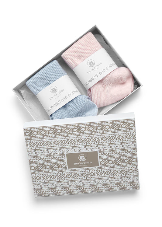 A gift set of two pairs of 100% cashmere socks in pastel blue and pink, presented in an open white box with a patterned lid. The socks are folded with white paper bands with the text 'THE CROFT HOUSE' and 'CASHMERE BED SOCKS'.