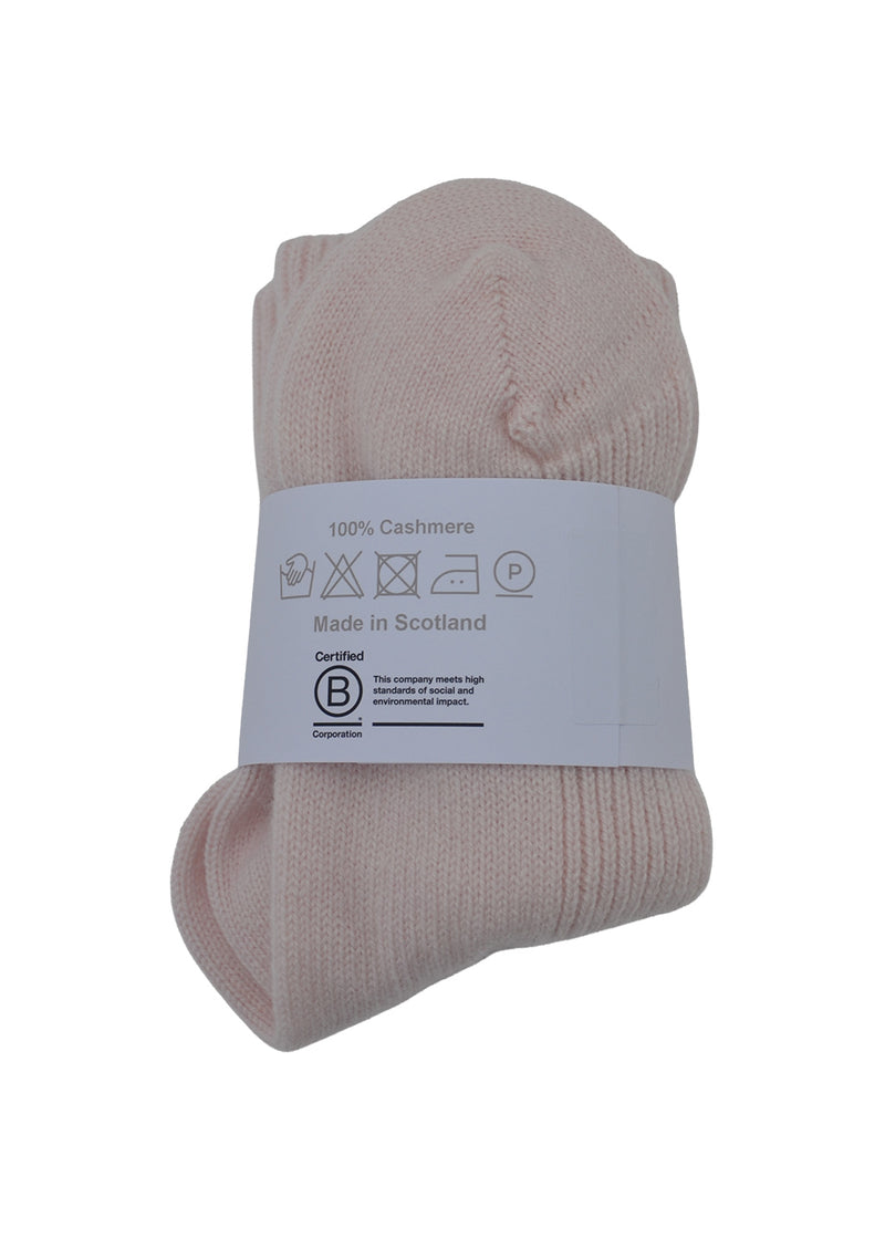 A pair of folded light pink cashmere bed socks with a white packaging band showing washing instructions and the B Corp logo, on a white background