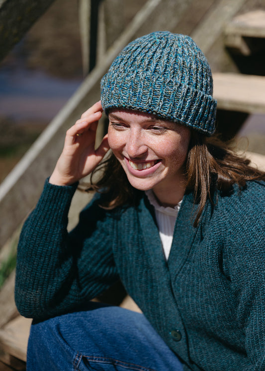 A woman smiling while wearing a teal blue and green marled knit beanie and a matching dark teal cardigan, sitting outdoors on wooden steps.