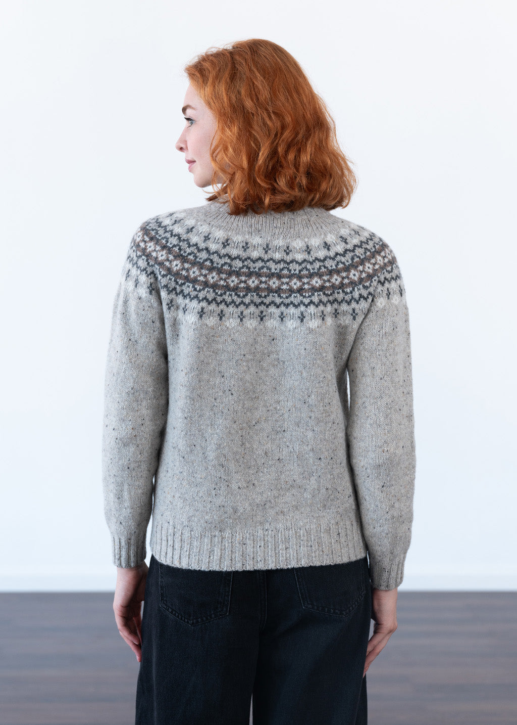 The back of a women with red hair wearing a grey fair isle jumper with pattern across the shoulders, with black jeans against a white studio wall
