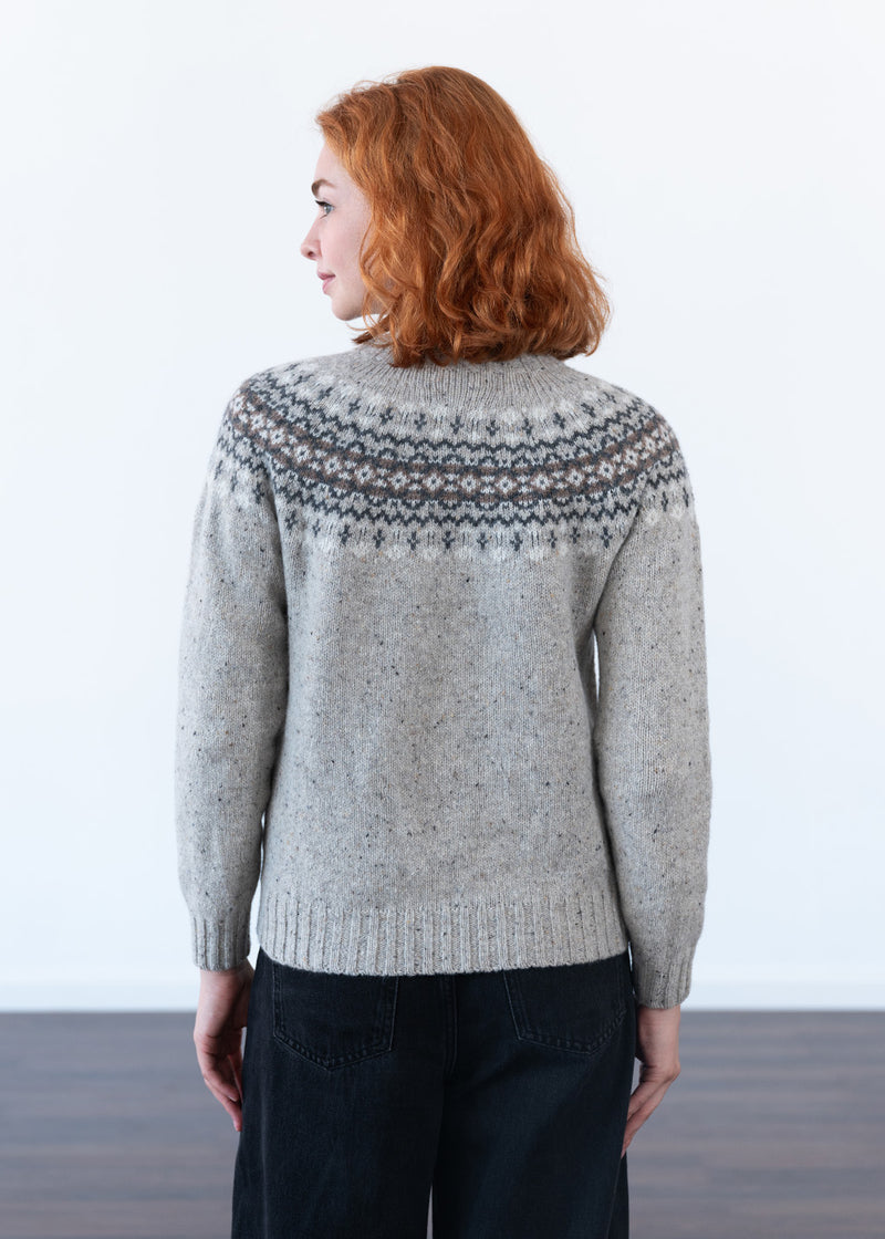 The back of a women with red hair wearing a grey fair isle jumper with pattern across the shoulders, with black jeans against a white studio wall