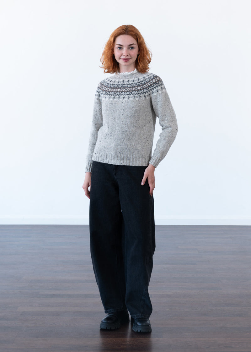 A full length image of a model with red hair wearing a grey fair isle jumper and wide leg black jeans with black boots against a white studio wall 