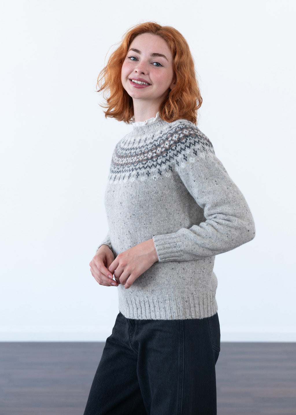A smiling model with red hair wearing a grey fair isle jumper with black jeans in a studio with white wall and dark wood floor