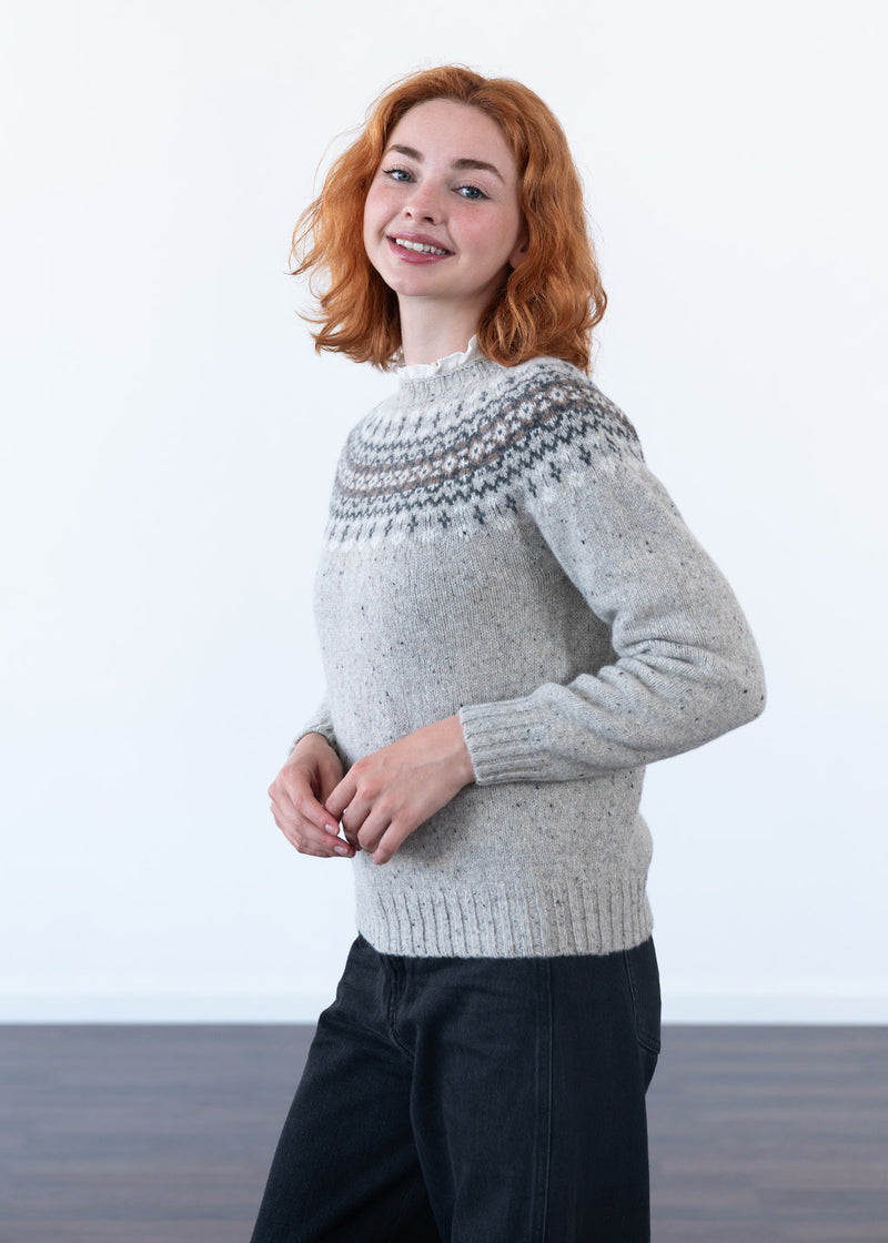 A smiling model with red hair wearing a grey fair isle jumper with black jeans in a studio with white wall and dark wood floor