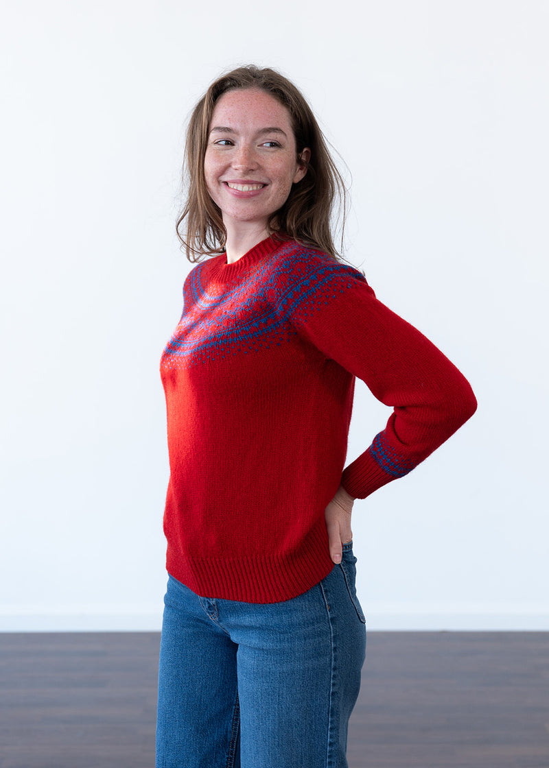 A woman wearing a bright red knit sweater with blue fair isle yoke details and blue jeans, standing against a plain white wall.