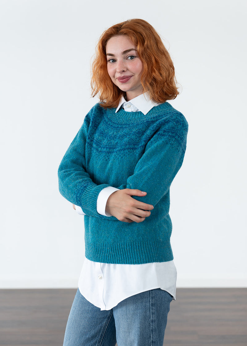 A woman with red hair wearing a teal blue fairisle knit jumper over a white collared shirt and blue jeans, standing against a plain white studio background.