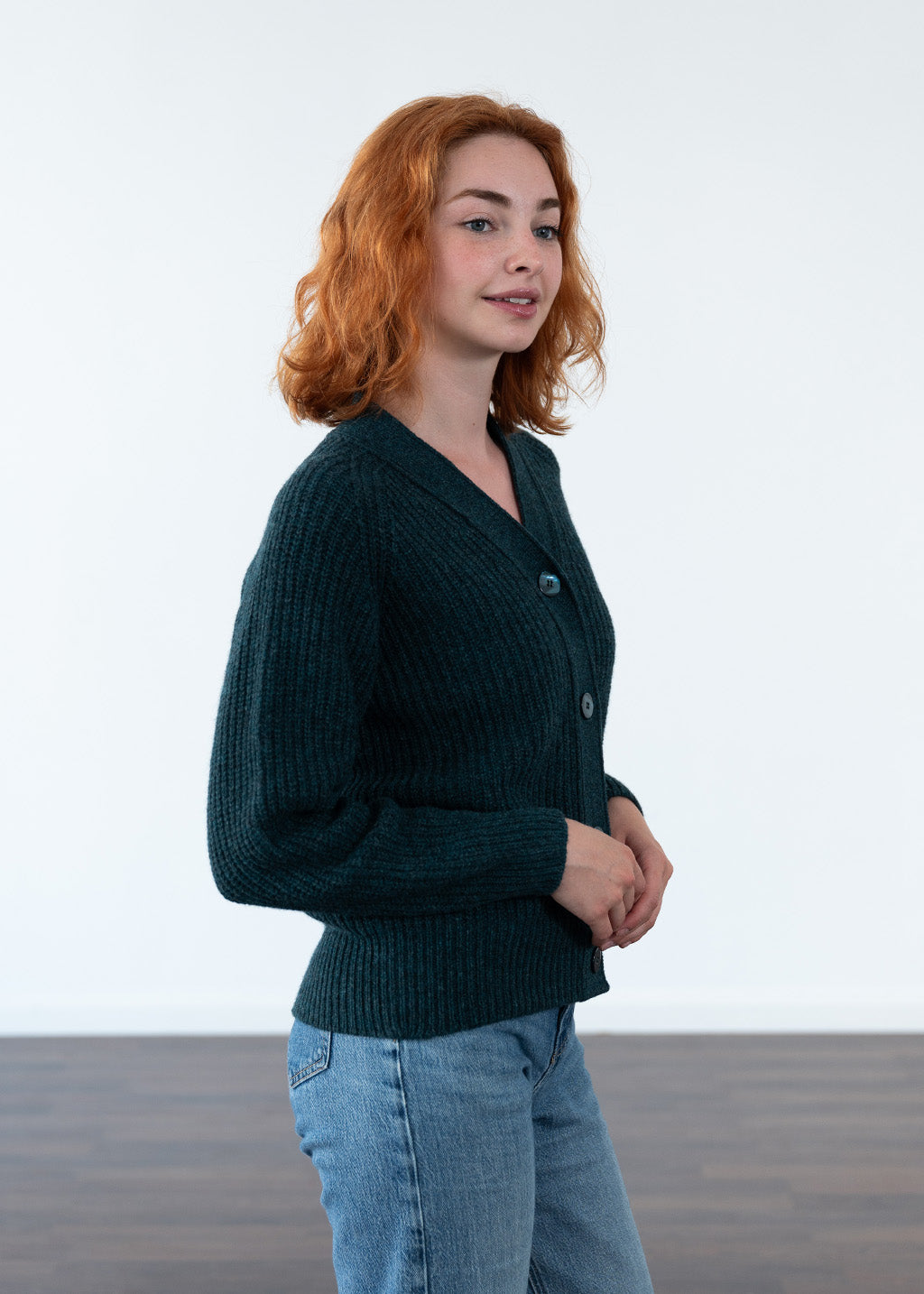A woman with red hair wearing a dark teal ribbed knit cardigan and light blue jeans, standing against a minimalist white wall with a wooden floor.