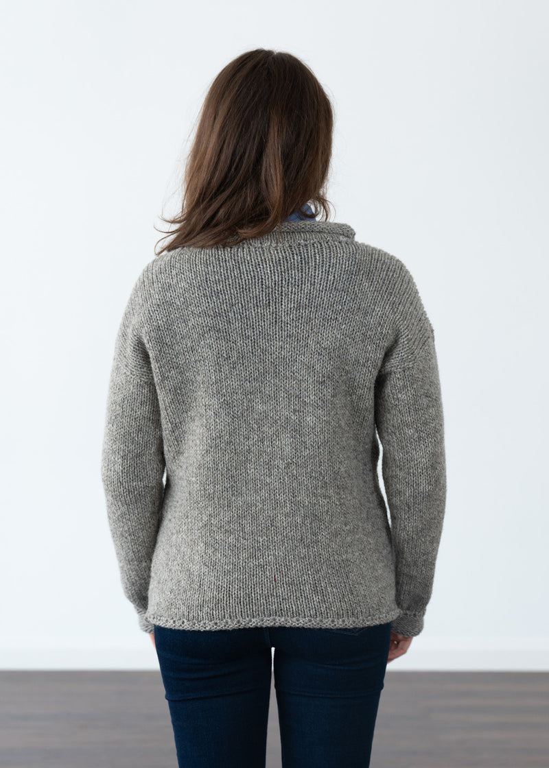 A woman viewed from behind wearing a marled grey-brown knit jumper and dark blue jeans, standing against a plain white wall.