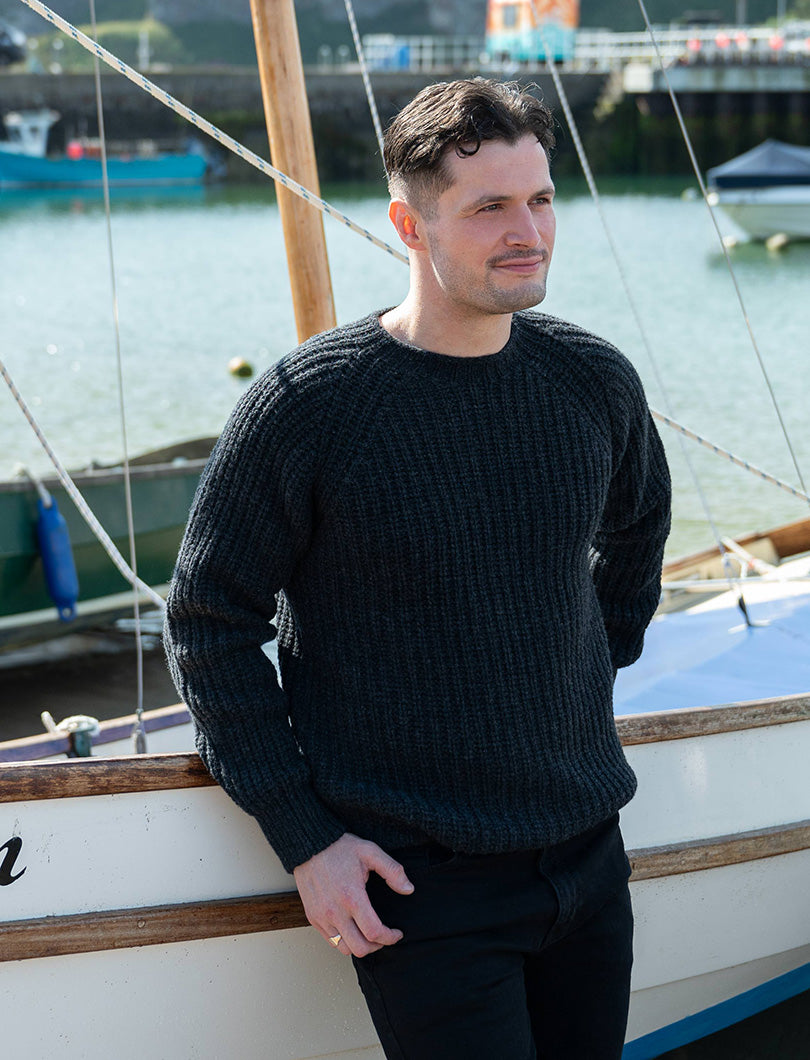Man wearing a charcoal grey ribbed sweater standing near a boat with water and boats in the background