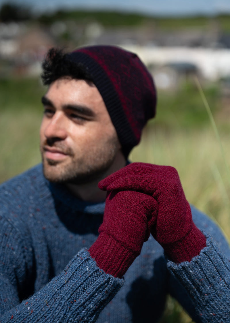 A man outdoors wearing burgundy knit lambs wool gloves, a dark burgundy and navy knit beanie and blue donegal wool jumper with a softly blurred natural background.
