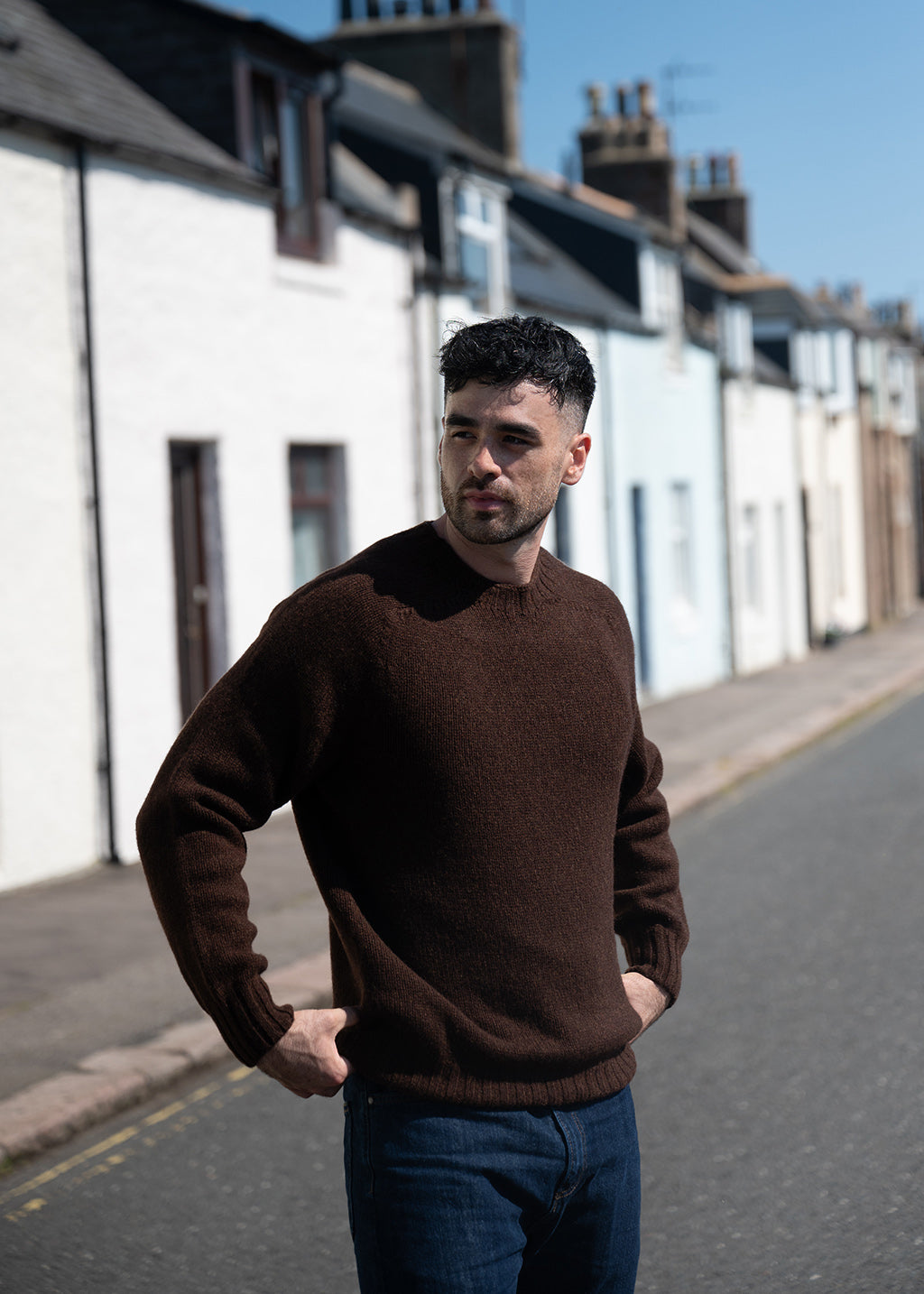 A man wearing a dark brown crewneck knit sweater and blue jeans standing on a street lined with white and blue houses.