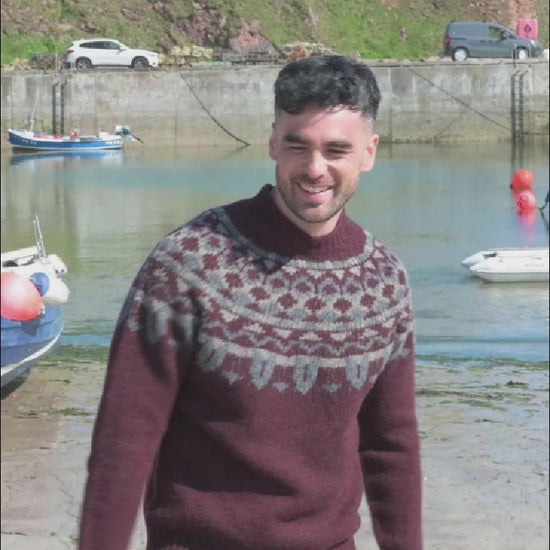 A video of a man by a small harbour wearing a chunky burgundy fair isle jumper with pattern around the yoke. 