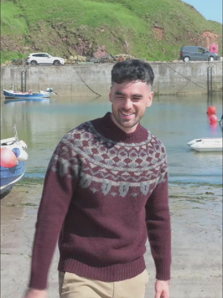 A video of a man by a small harbour wearing a chunky burgundy fair isle jumper with pattern around the yoke. 