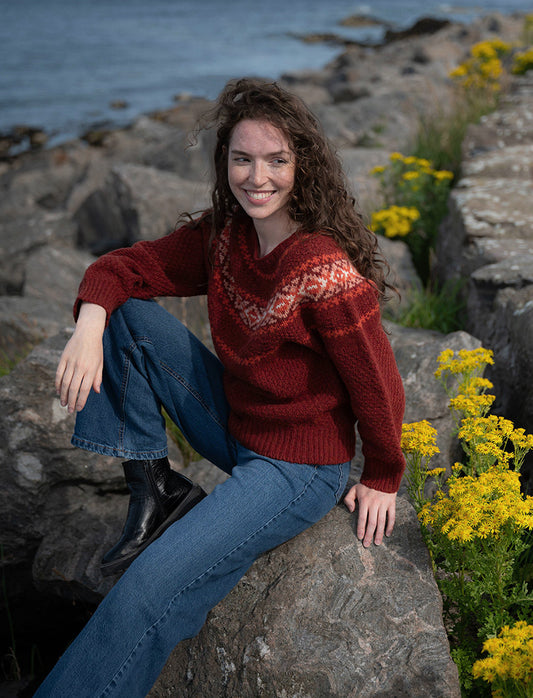 Girl wearing a russet and orange fair isle textured jumper whilst sitting on rocks by the sea, surrounded by yellow flowers