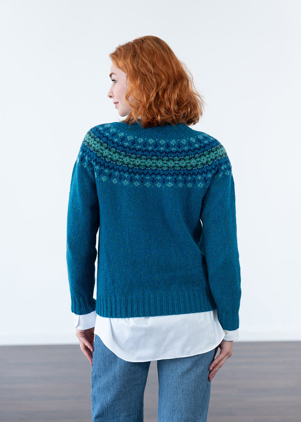 A woman with red hair is shown from the back wearing a teal blue Fair Isle knit sweater over a white shirt and blue jeans, set against a clean white studio background.