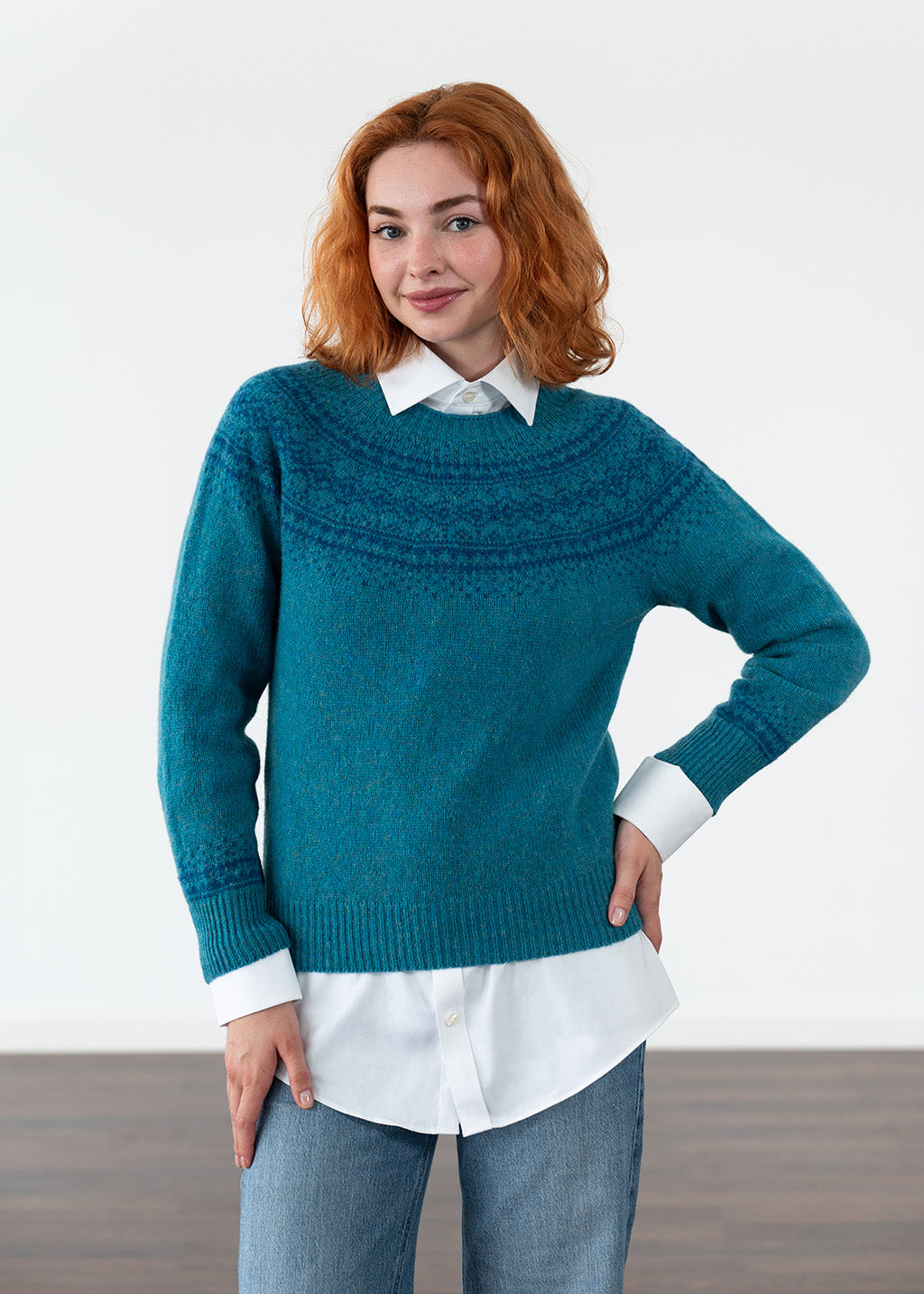 A woman with red hair wearing a teal blue knit sweater with a Fair Isle pattern across the chest, layered over a white collared shirt and blue jeans, standing against a white studio background.