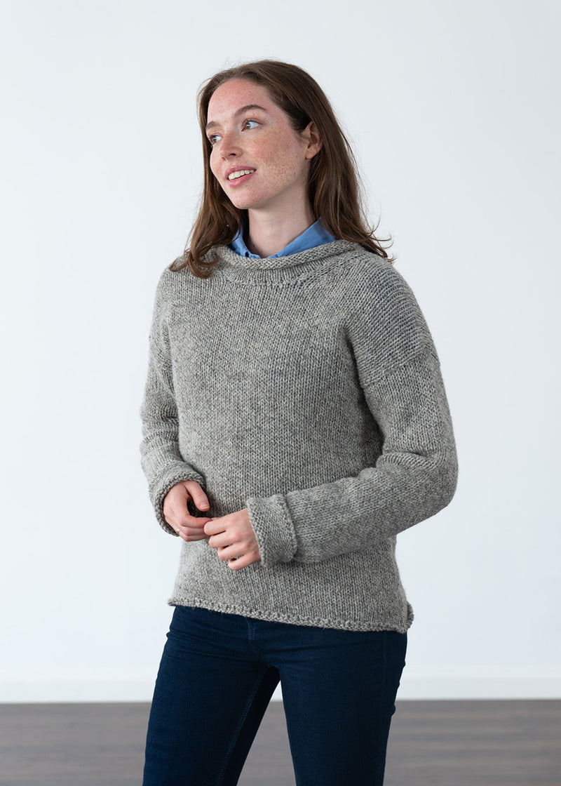 A woman modeling a chunky knit sweater in a heathered gray color, layered over a light blue collared shirt and paired with dark blue jeans, set against a plain white studio background.