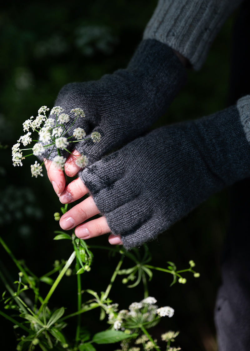 A close-up of a person's hands wearing dark grey knit cashmere fingerless gloves, holding small white wildflowers against a dark, blurred outdoor background.