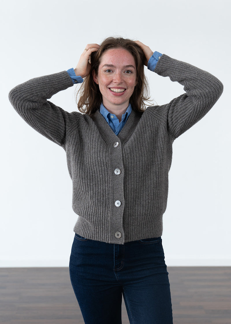A woman wearing a brown ribbed button-up cardigan over a blue collared shirt and dark jeans, standing against a plain white studio background with her hands in her hair.