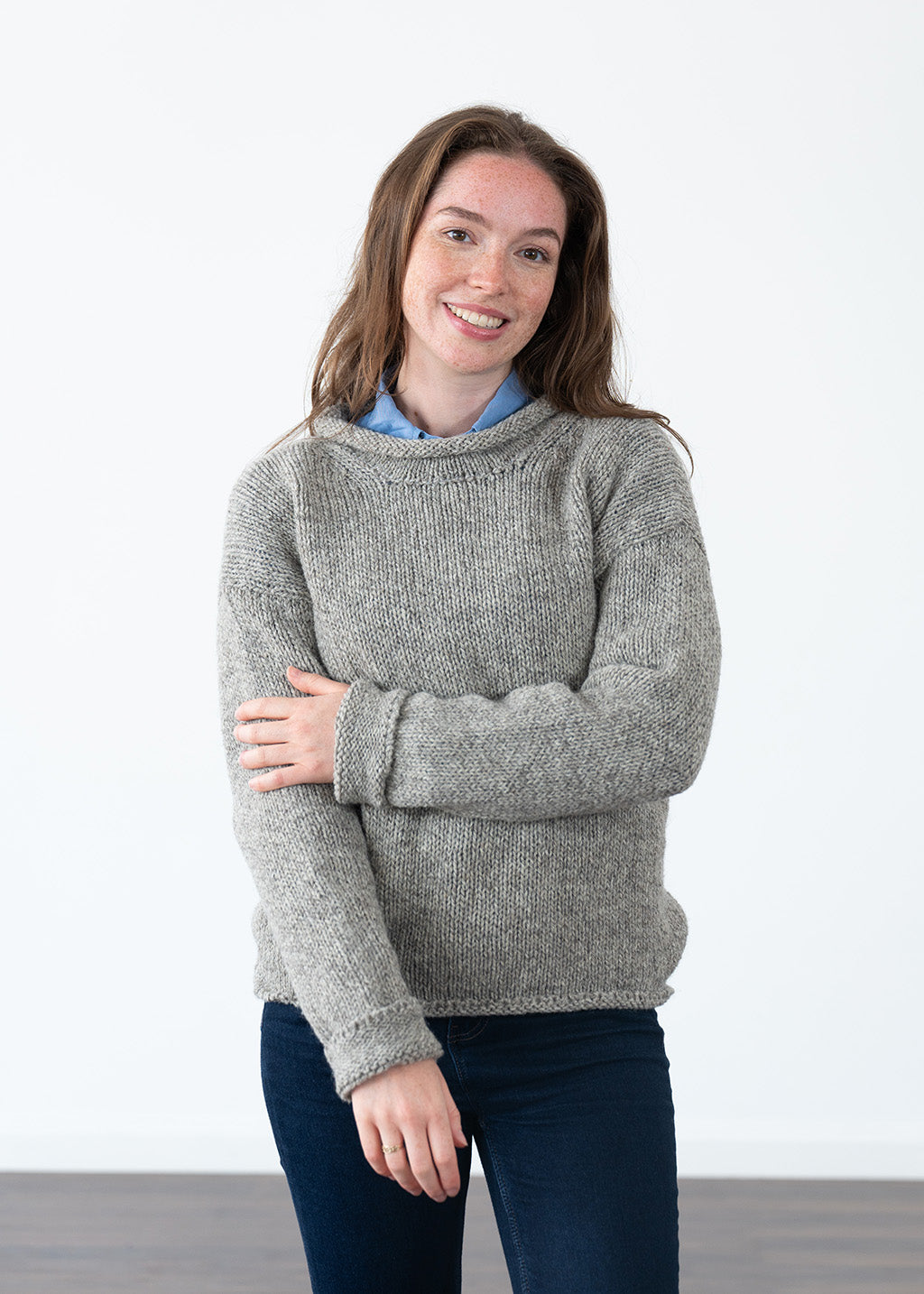 A woman wearing a chunky knit grey jumper over a light blue collared shirt and dark blue jeans, standing against a plain white wall.
