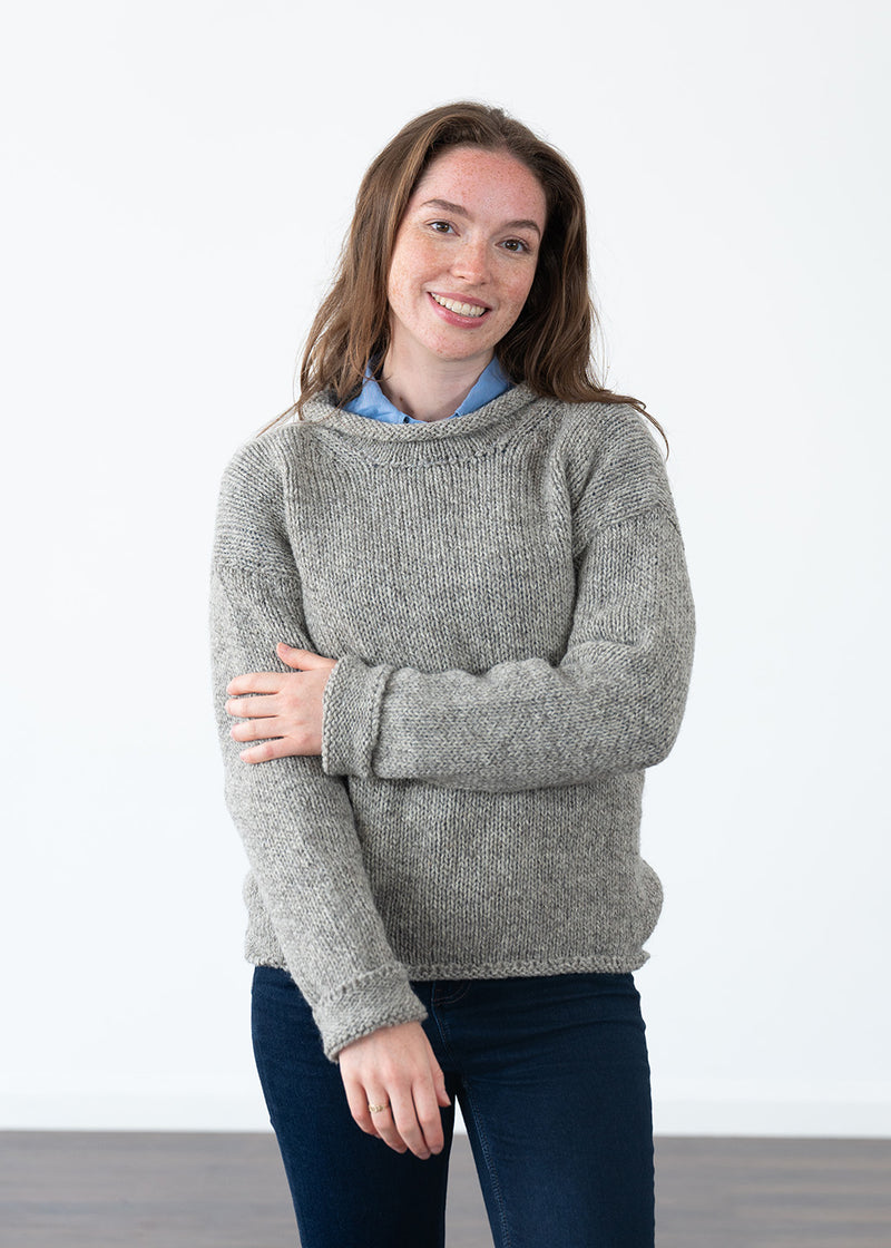 A woman wearing a chunky knit grey jumper over a light blue collared shirt and dark blue jeans, standing against a plain white wall.