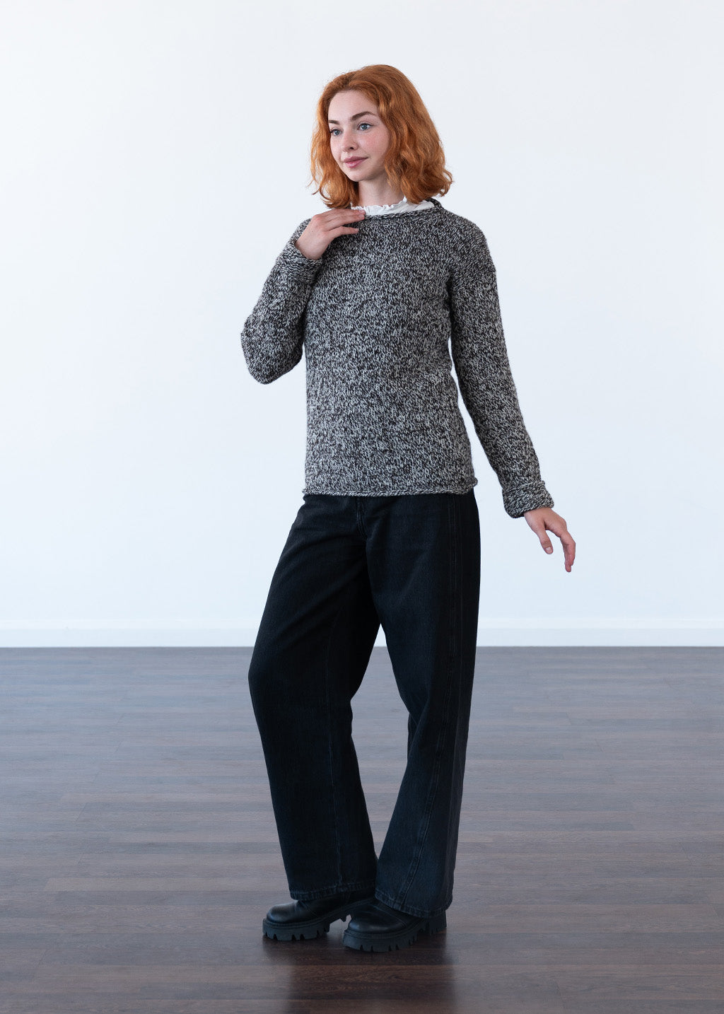 A woman with red hair stands in a studio wearing an ecru and black marled knit sweater, black wide-leg jeans, and black chunky soled boots against a white wall and dark wood floor.