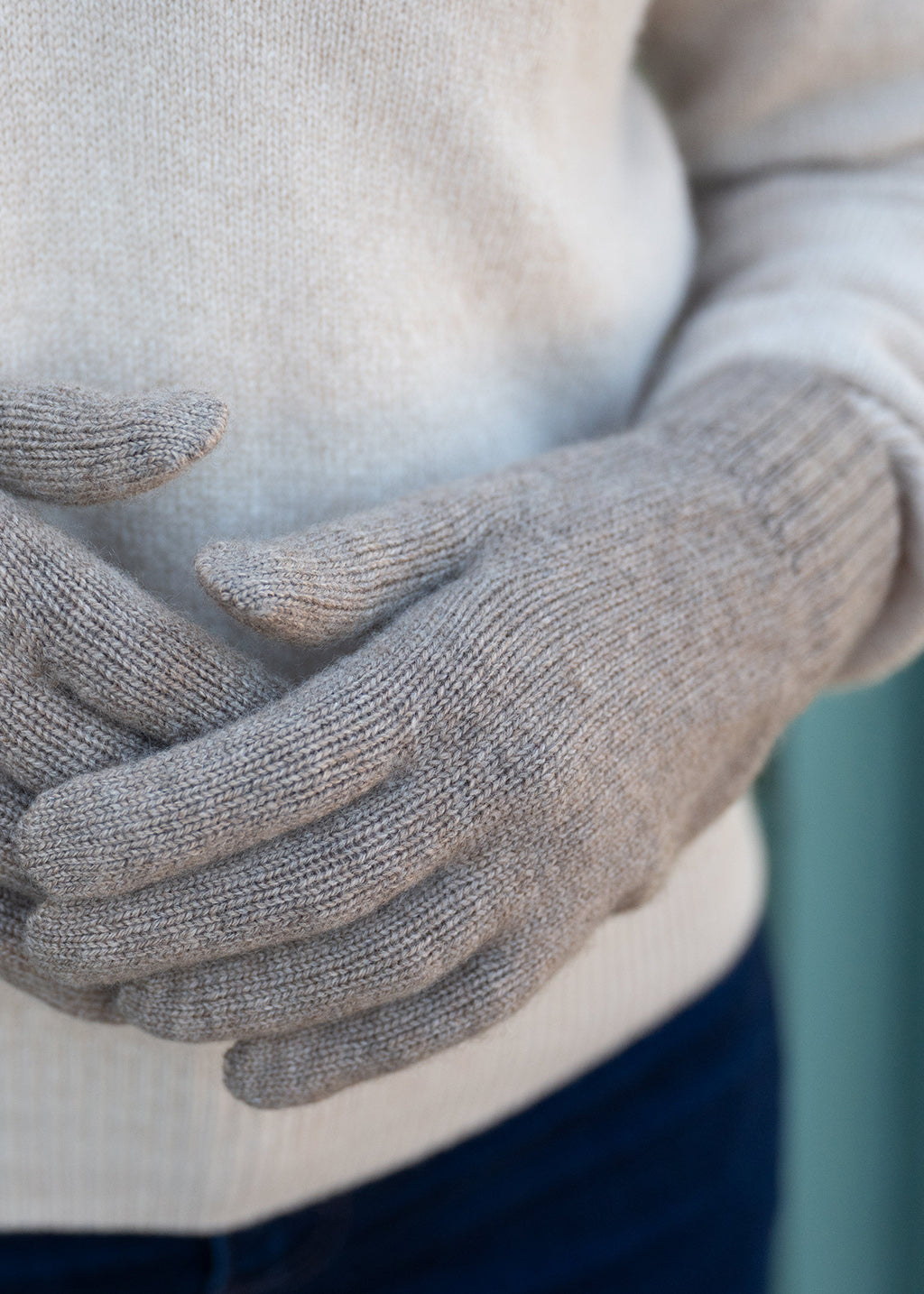 Close-up of a women wearing light brown knit cashmere gloves over a oatmeal sweater and jeans.