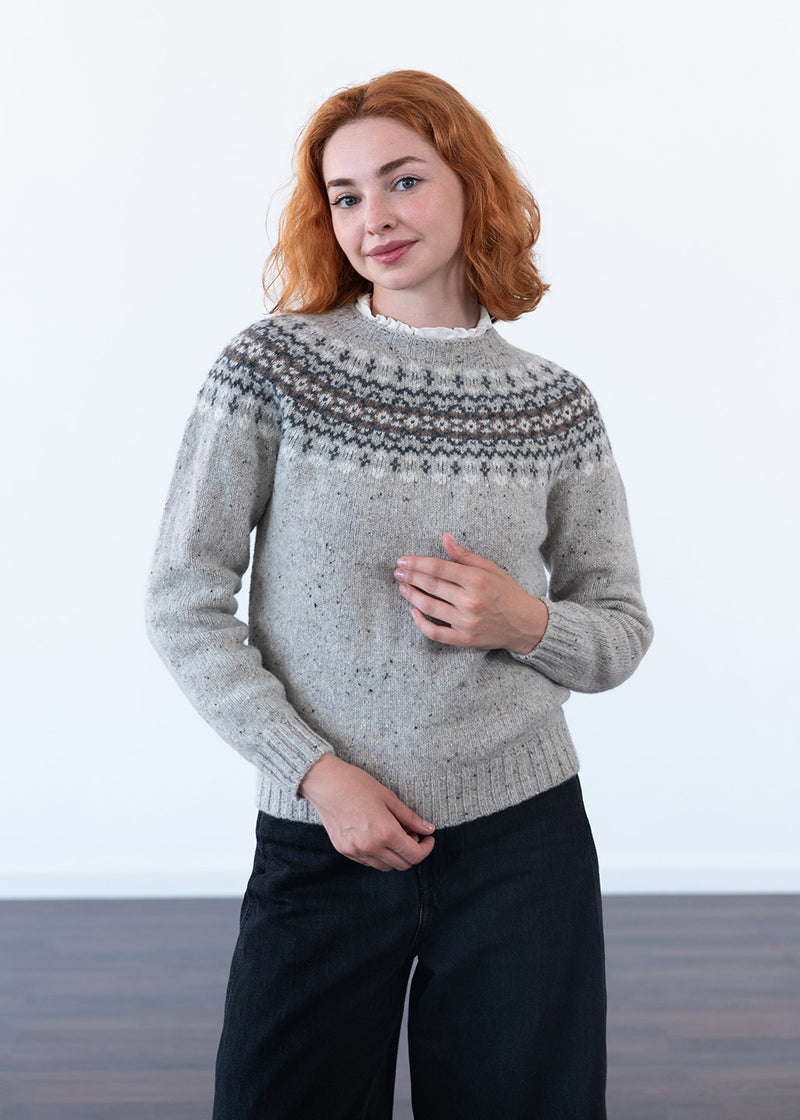 A women with red hair wearing a grey fair isle jumper with small ruffled blouse collar on show, and black jeans against a white studio wall