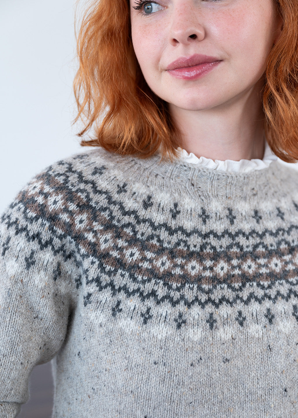 A close up of a women with red hair wearing a fair isle jumper in grey donegal wool with pattern across the chest and shoulders in brown and dark grey