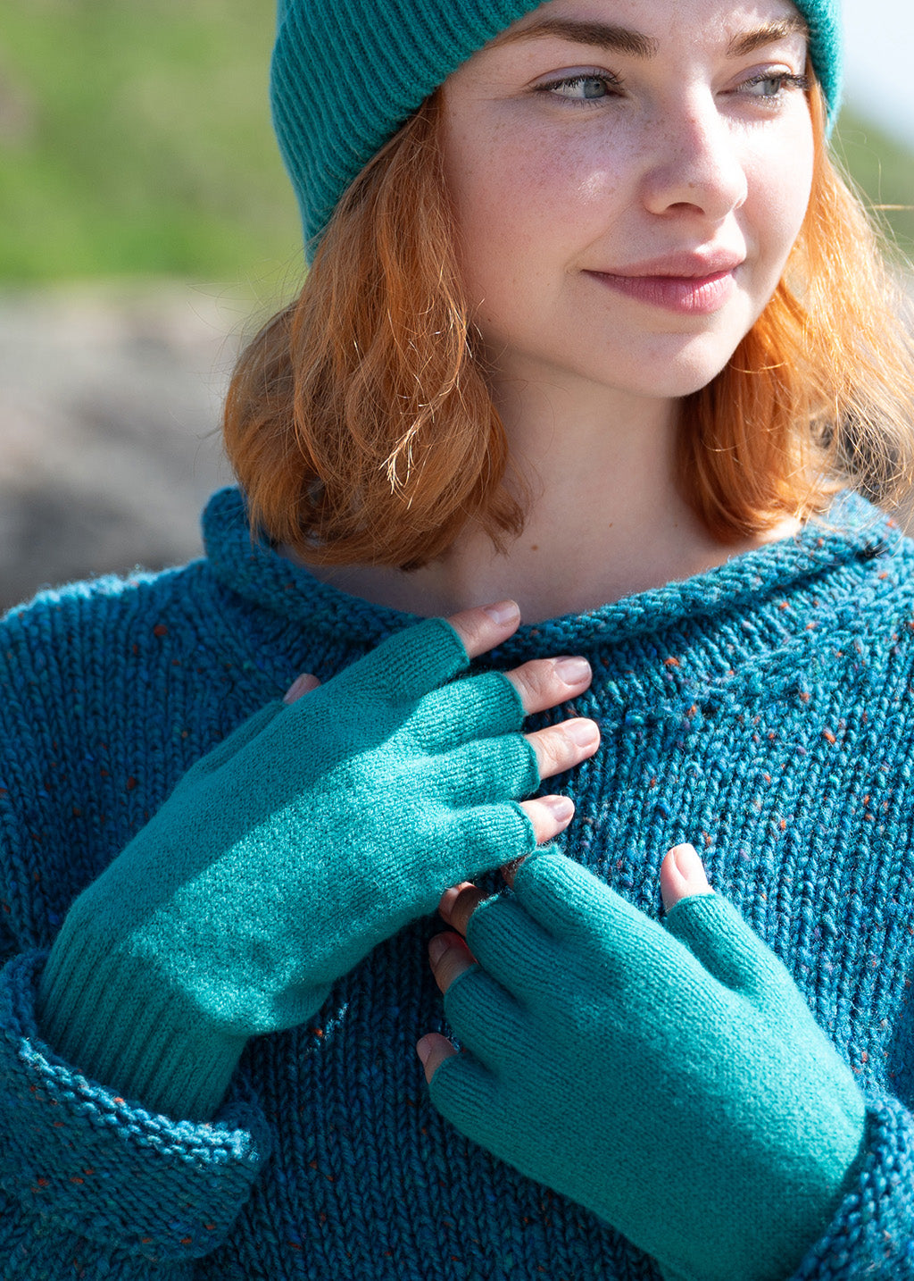A close-up of a woman wearing sea green knit fingerless gloves, matching ribbed beanie hat and a chunky teal donegal wool sweater. She is looking off-camera with a soft smile, set against a blurred outdoor background.