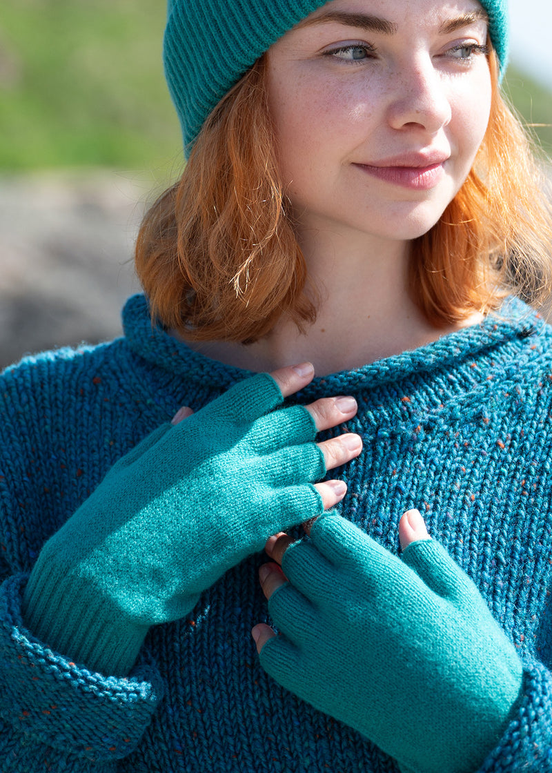 A close-up of a woman wearing sea green knit fingerless gloves, matching ribbed beanie hat and a chunky teal donegal wool sweater. She is looking off-camera with a soft smile, set against a blurred outdoor background.