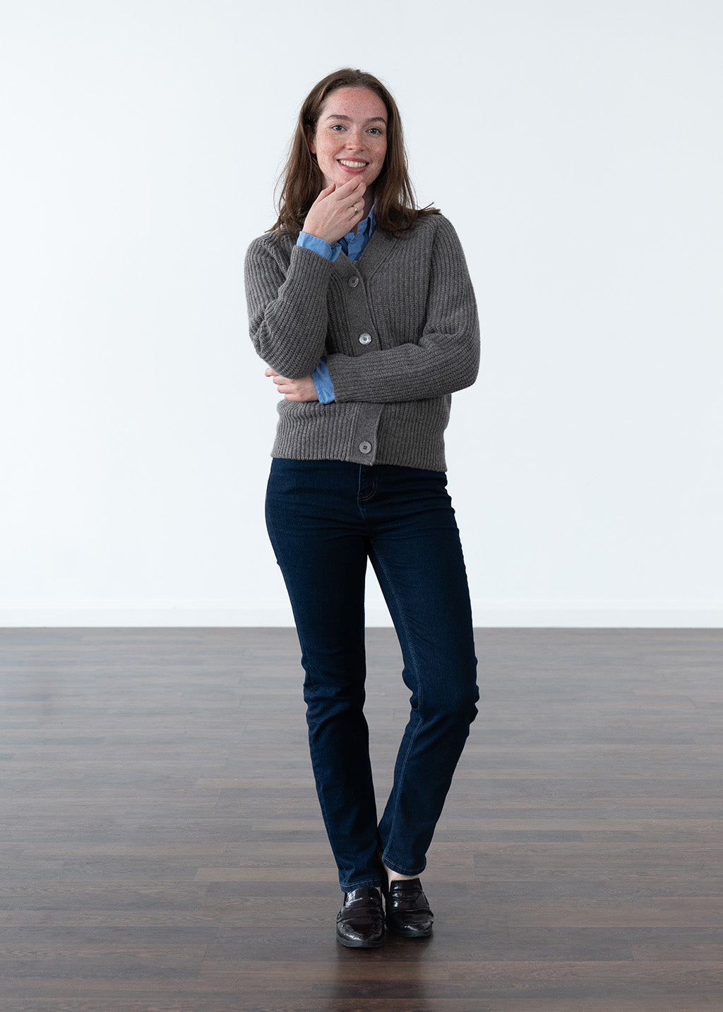 A woman stands in a studio wearing a grey-brown ribbed cardigan over a blue button-up shirt, paired with dark navy jeans and black shoes.