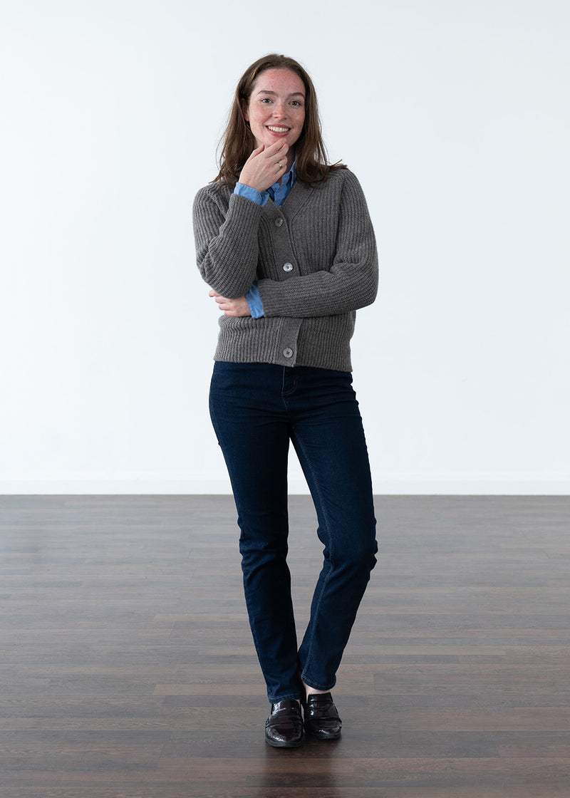 A woman stands in a studio wearing a grey-brown ribbed cardigan over a blue button-up shirt, paired with dark navy jeans and black shoes.