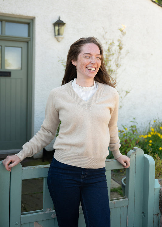 A woman laughing while wearing a beige v-neck jumper over a white ruffled blouse and dark blue jeans, standing outdoors in front of a white stucco wall and a sage green wooden gate.