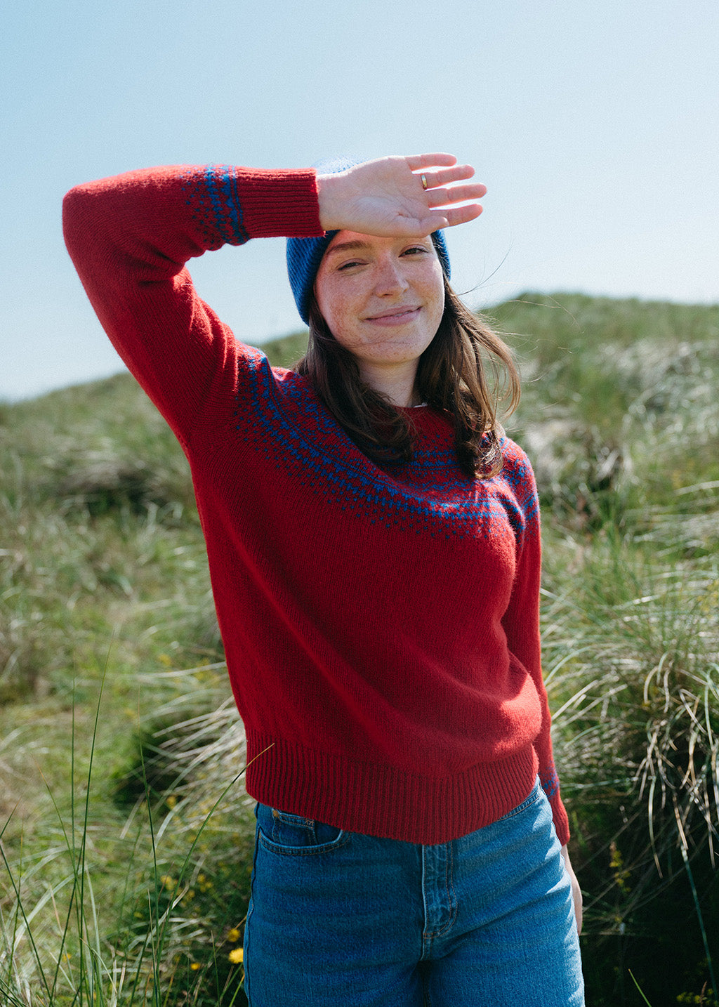 A woman wearing a red knit jumper with blue Fair Isle yoke details, blue jeans, and a blue beanie stands outdoors in a grassy field under a clear blue sky, shielding her eyes from the sun.