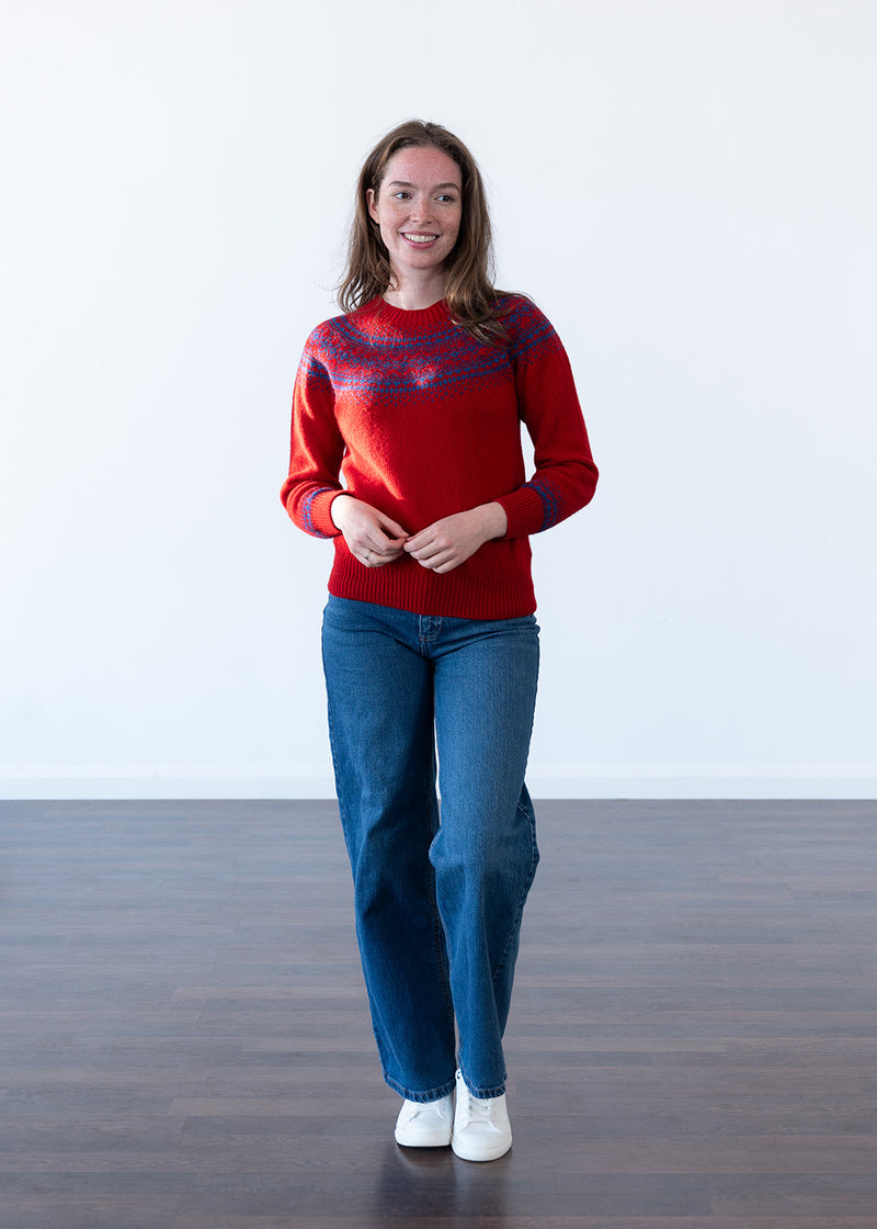 A woman modeling a red knit jumper with a blue fair isle pattern across the chest and shoulders, paired with blue jeans and white trainers, standing against a white wall and dark wood floor.